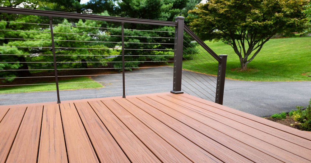 The image shows a newly constructed outdoor deck with composite decking material in a warm brown color. The deck features a sleek, modern cable railing system with horizontal metal cables and dark metal posts.